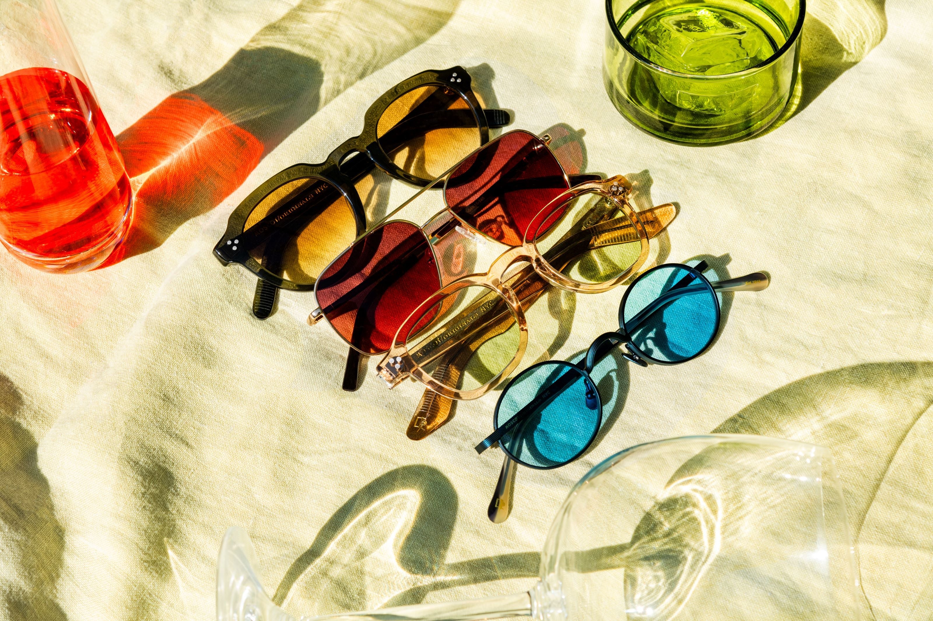 Four pairs of MOSCOT sunglasses with different lens colors (blue, red, brown) on a light fabric. Nearby, red and green glasses cast colorful shadows, creating a vibrant, summery scene.